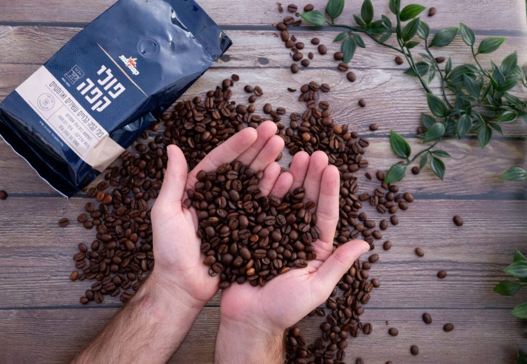 פולי קפה טריים בידיים מעל שולחן עץ, ליד שקית קפה של קפה עלית – בלנד מקומי שנרקח במיוחד לטעם הישראלי, כמו זה שאתם רגילים לשתות גם מחוץ לבית.